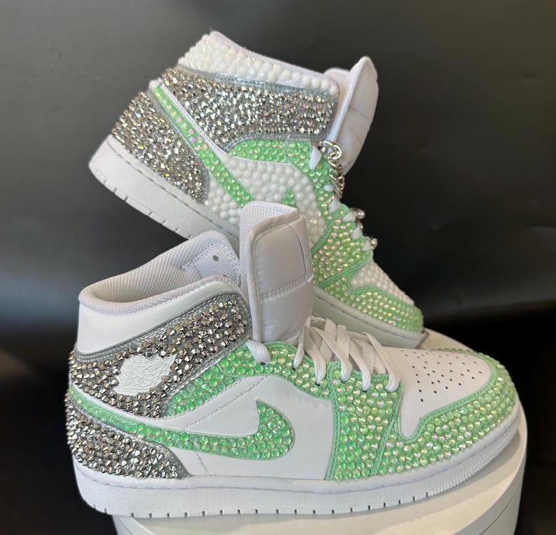 May include: White high-top sneakers with green and silver rhinestones. The sneakers have a white swoosh logo and a white sole.