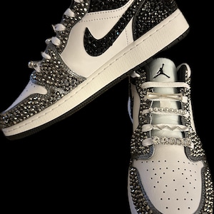 Puede incluir: Un par de zapatillas blancas de ca&ntilde;a baja con detalles negros y un logotipo Swoosh negro. Las zapatillas est&aacute;n cubiertas de pedrer&iacute;a y tienen un cord&oacute;n blanco con detalles plateados y perlados.