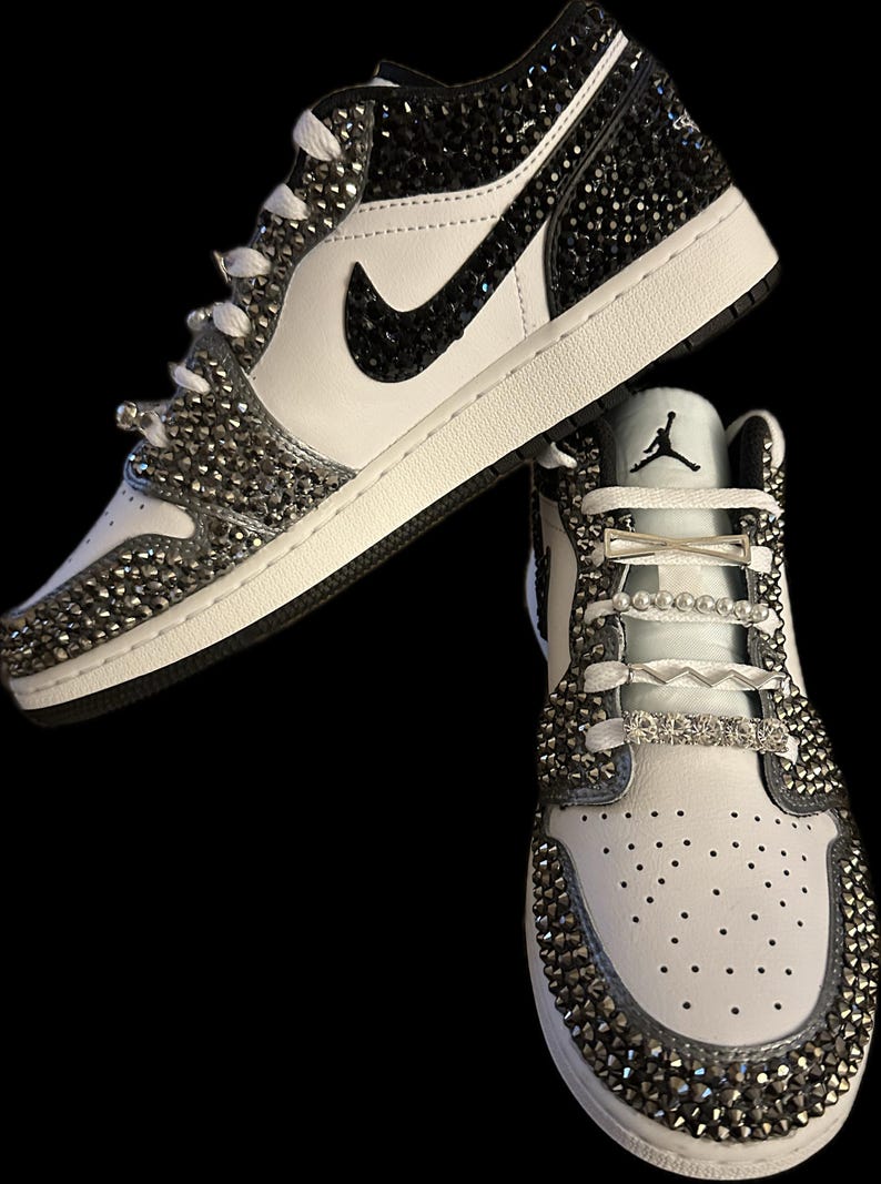 Puede incluir: Un par de zapatillas blancas de ca&ntilde;a baja con detalles negros y un logotipo Swoosh negro. Las zapatillas est&aacute;n cubiertas de pedrer&iacute;a y tienen un cord&oacute;n blanco con detalles plateados y perlados.