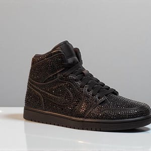 May include: Black high-top sneaker covered in small, sparkling embellishments. The shoe features a black sole, laces, and the iconic swoosh design. The sneaker is set against a neutral background, highlighting its details.