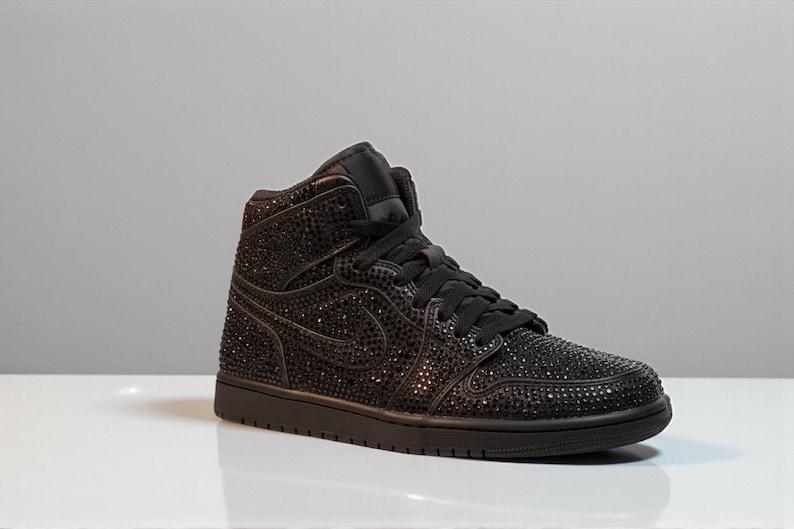May include: Black high-top sneaker covered in small, sparkling embellishments. The shoe features a black sole, laces, and the iconic swoosh design. The sneaker is set against a neutral background, highlighting its details.