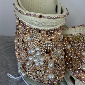 Pu&ograve; includere: Zoccoli bianchi decorati con perle, strass e dettagli dorati. Gli zoccoli presentano un design a farfalla e un motivo floreale.