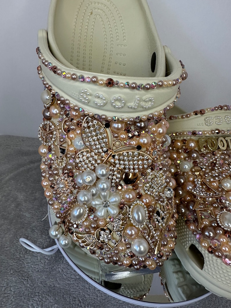 Pu&ograve; includere: Zoccoli bianchi decorati con perle, strass e dettagli dorati. Gli zoccoli presentano un design a farfalla e un motivo floreale.