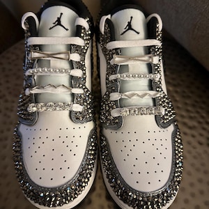 Puede incluir: Zapatillas blancas de cuero con pedrer&iacute;a plateada y abalorios de zapatos plateados. Las zapatillas tienen un logotipo Swoosh negro y cordones blancos.