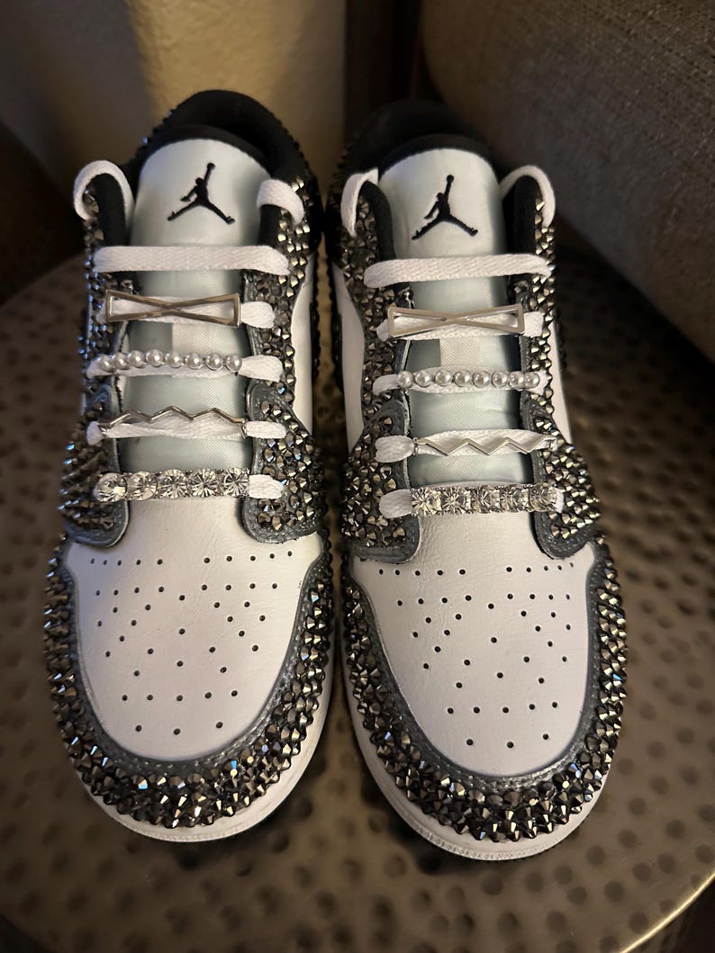 Puede incluir: Zapatillas blancas de cuero con pedrer&iacute;a plateada y abalorios de zapatos plateados. Las zapatillas tienen un logotipo Swoosh negro y cordones blancos.