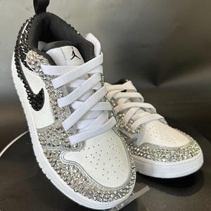 Puede incluir: Zapatillas blancas de cuero con detalles negros y un dise&ntilde;o de cristal. Las zapatillas tienen un cierre de cordones blanco y un logotipo swoosh negro.
