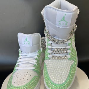 May include: White high-top sneakers with green accents and rhinestones. The sneakers have a silver chain detail and the word "AIR" is written on the side.