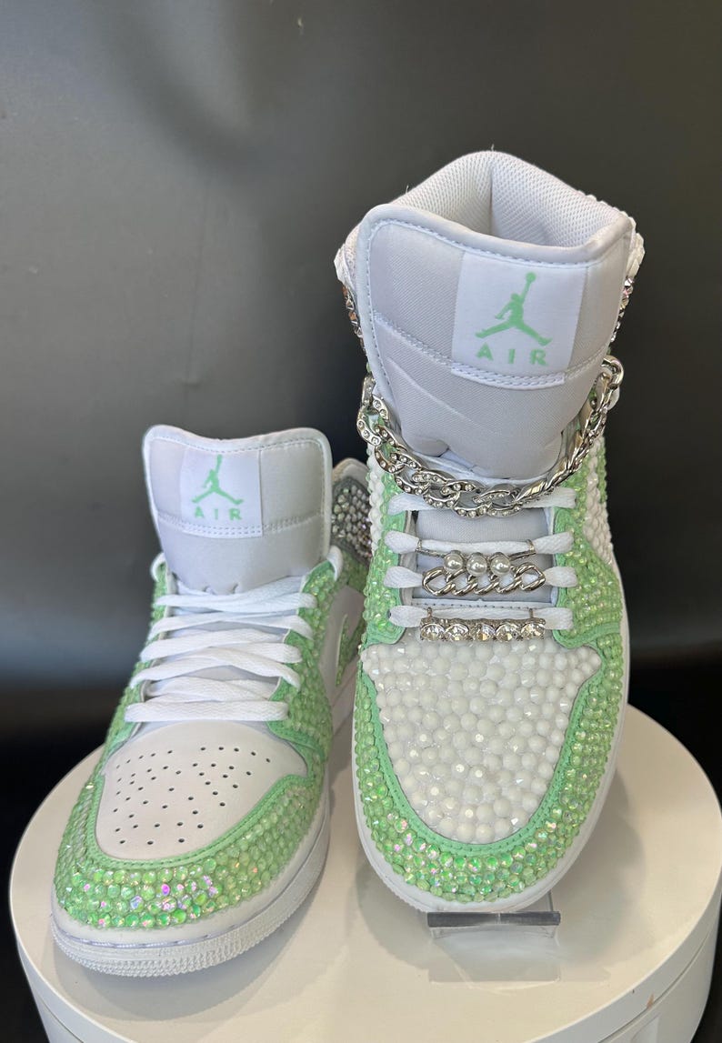 May include: White high-top sneakers with green accents and rhinestones. The sneakers have a silver chain detail and the word "AIR" is written on the side.