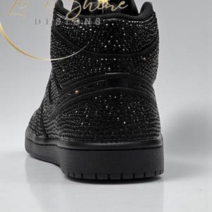 May include: Black high-top sneaker covered in sparkling embellishments. The shoe features a solid black sole and a textured upper, with the words "Let it Shine Designs" in gold script.