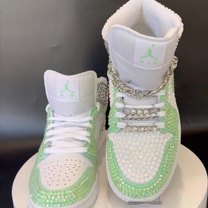 May include: A pair of white high-top sneakers with green rhinestones and a silver chain detail. The sneakers have the word "AIR" on the side.