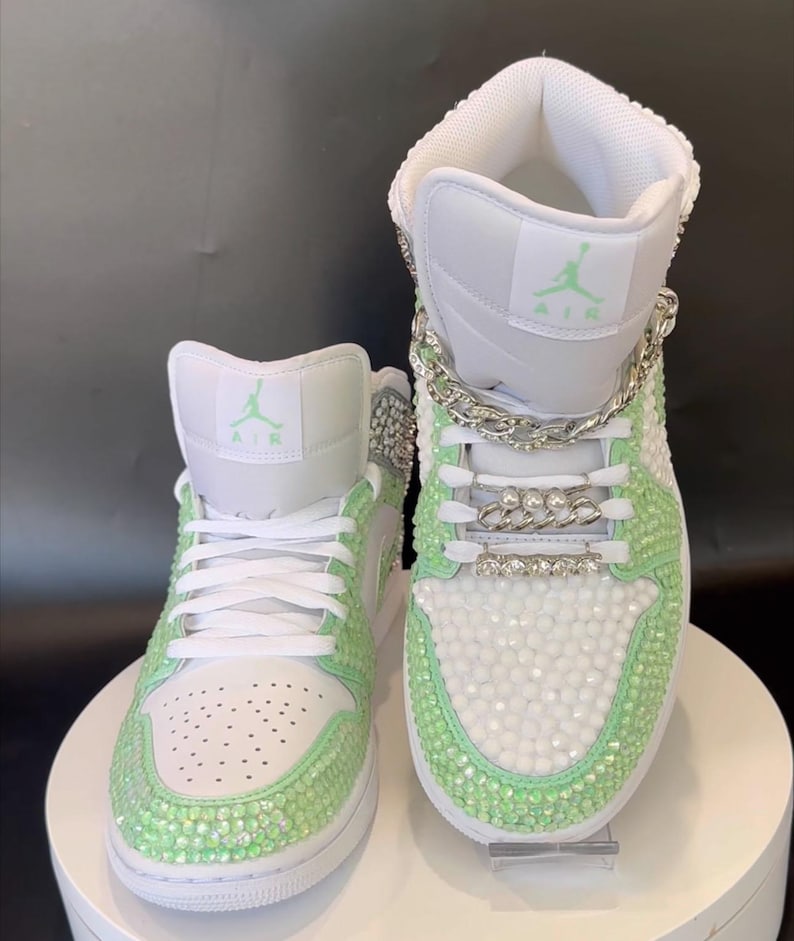 May include: A pair of white high-top sneakers with green rhinestones and a silver chain detail. The sneakers have the word "AIR" on the side.