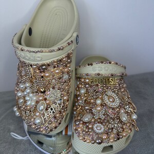 Pu&ograve; includere: Un paio di zoccoli beige decorati con perle, strass e dettagli dorati. Gli zoccoli hanno la scritta "FOOTSIE" su di loro.