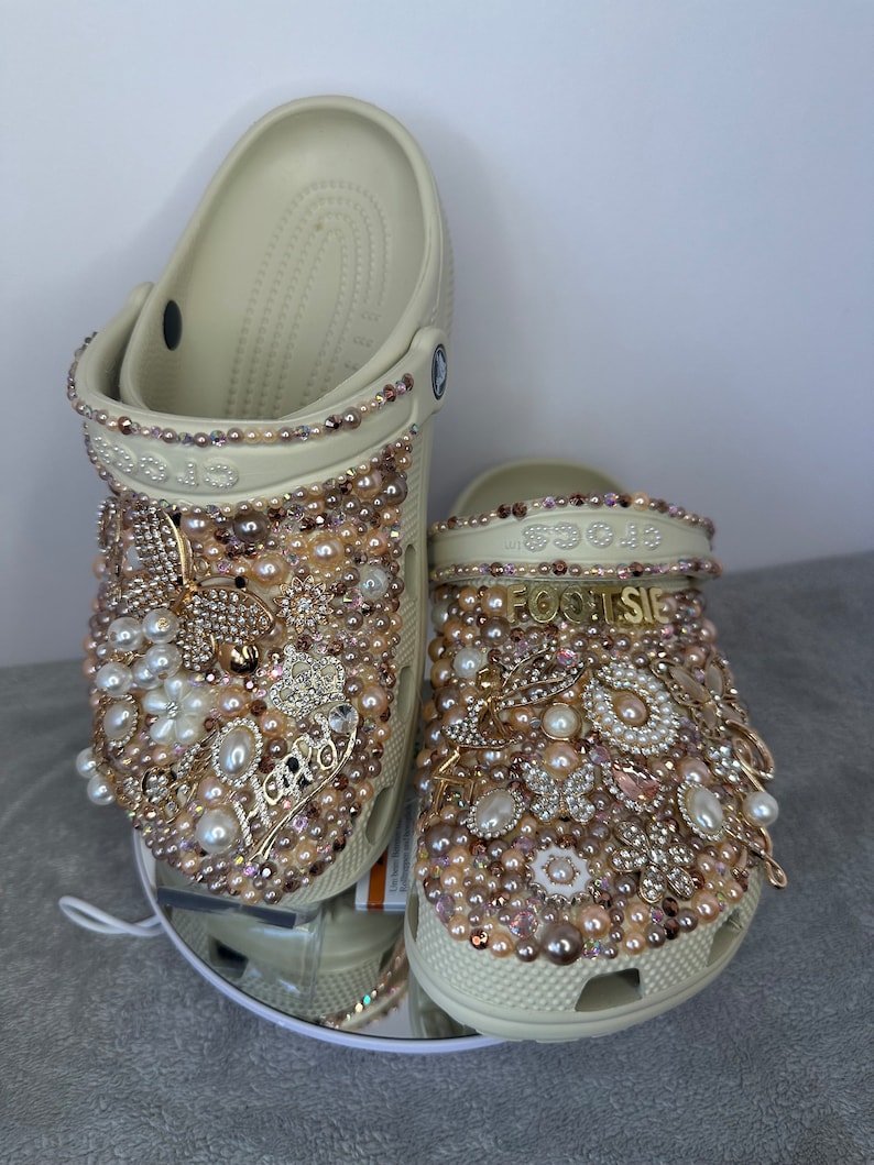 Pu&ograve; includere: Un paio di zoccoli beige decorati con perle, strass e dettagli dorati. Gli zoccoli hanno la scritta "FOOTSIE" su di loro.