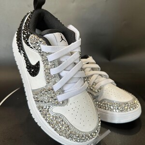 Puede incluir: Zapatillas bajas de cuero blanco con detalles negros y un dise&ntilde;o cubierto de cristales. Las zapatillas tienen un cierre de cordones blanco y un logotipo Swoosh negro.