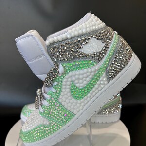 May include: White high-top sneakers with a green and silver rhinestone design. The sneakers have a white swoosh logo and a white sole.