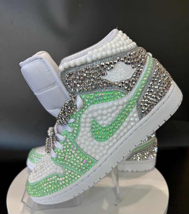 May include: White high-top sneakers with a green and silver rhinestone design. The sneakers have a white swoosh logo and a white sole.