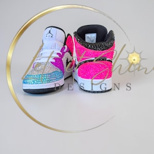 May include: A pair of bedazzled high-top sneakers. The sneakers feature a white tongue, a black collar, and a vibrant color scheme of pink, blue, and silver rhinestones. The iconic Air Jordan logo is visible on the tongue.