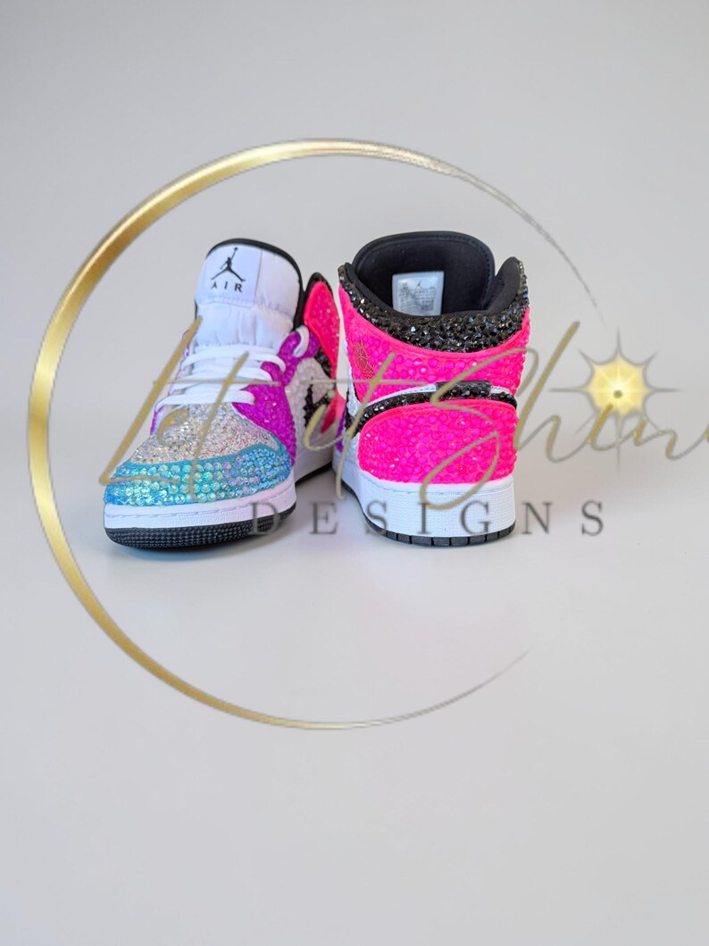 May include: A pair of bedazzled high-top sneakers. The sneakers feature a white tongue, a black collar, and a vibrant color scheme of pink, blue, and silver rhinestones. The iconic Air Jordan logo is visible on the tongue.