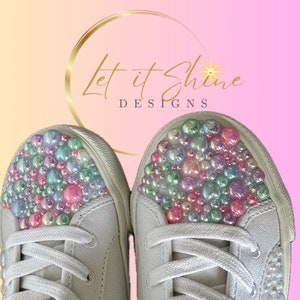 May include: White canvas sneakers with iridescent pearl embellishments on the toe. The shoes have white laces and are decorated with a cluster of pearls.