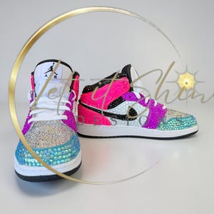 May include: A pair of bedazzled sneakers in a vibrant color scheme. The shoes feature a white base with accents of pink, purple, and teal, and are covered in sparkling rhinestones. The iconic Jordan logo is visible on the tongue.