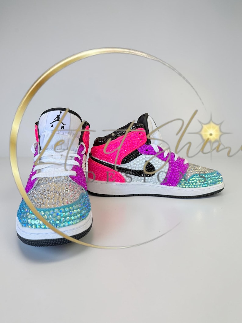 May include: A pair of bedazzled sneakers in a vibrant color scheme. The shoes feature a white base with accents of pink, purple, and teal, and are covered in sparkling rhinestones. The iconic Jordan logo is visible on the tongue.