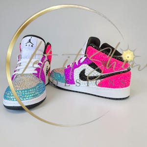 May include: A pair of bedazzled high-top sneakers. The shoes feature a white base with black accents, and are embellished with iridescent rhinestones in shades of blue, pink, and purple. The iconic Air Jordan logo is visible on the tongue.