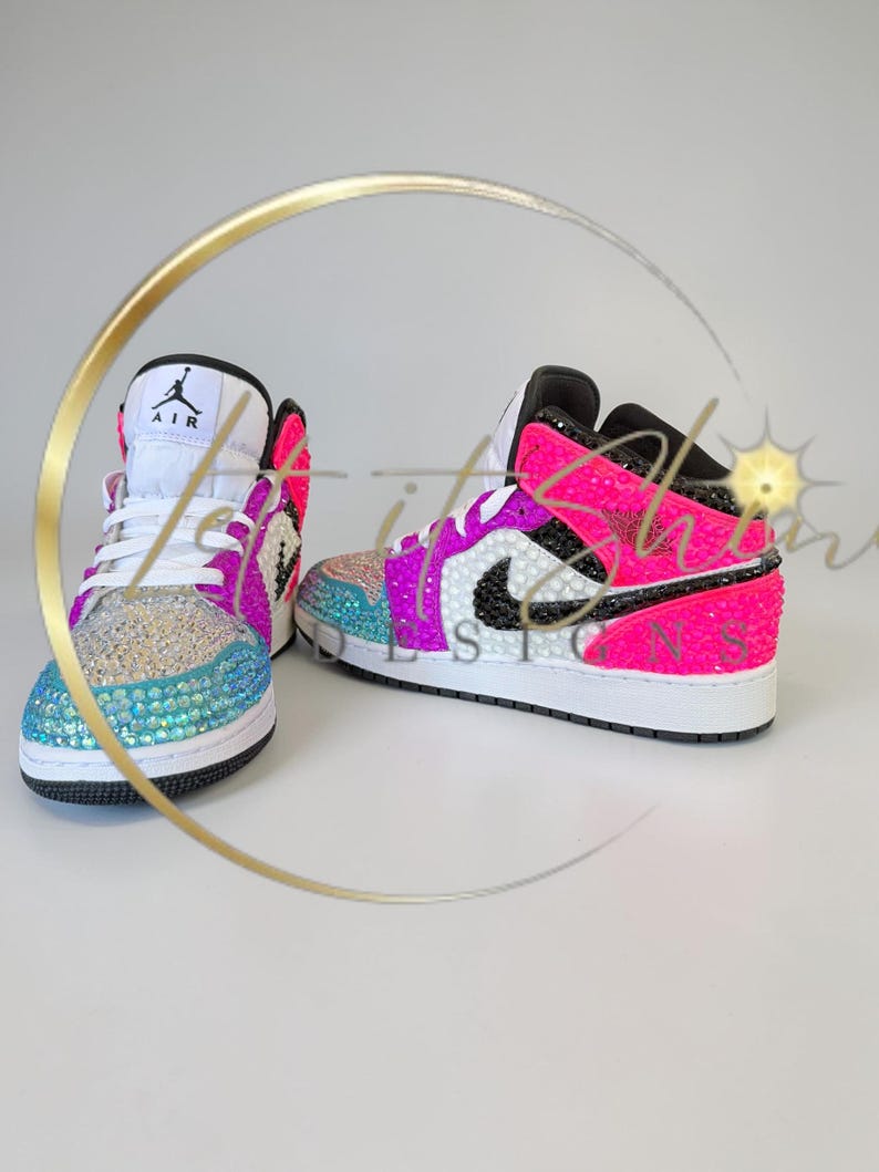May include: A pair of bedazzled high-top sneakers. The shoes feature a white base with black accents, and are embellished with iridescent rhinestones in shades of blue, pink, and purple. The iconic Air Jordan logo is visible on the tongue.
