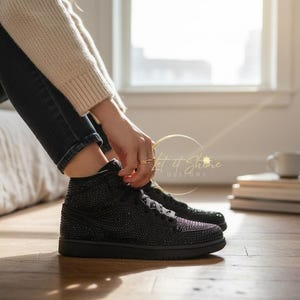 May include: Black high-top sneakers with sparkling embellishments. The shoes have black laces and soles. The image shows the sneakers on a wooden floor, with a person's legs and a bed in the background.