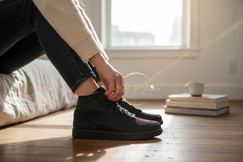 May include: Black high-top sneakers with sparkling embellishments. The shoes have black laces and soles. The image shows the sneakers on a wooden floor, with a person's legs and a bed in the background.
