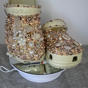 Pu&ograve; includere: Un paio di zoccoli bianchi decorati con perle, strass e dettagli dorati. Gli zoccoli hanno un design floreale e la parola "crocs" &egrave; visibile sulla parte superiore di uno zoccolo.