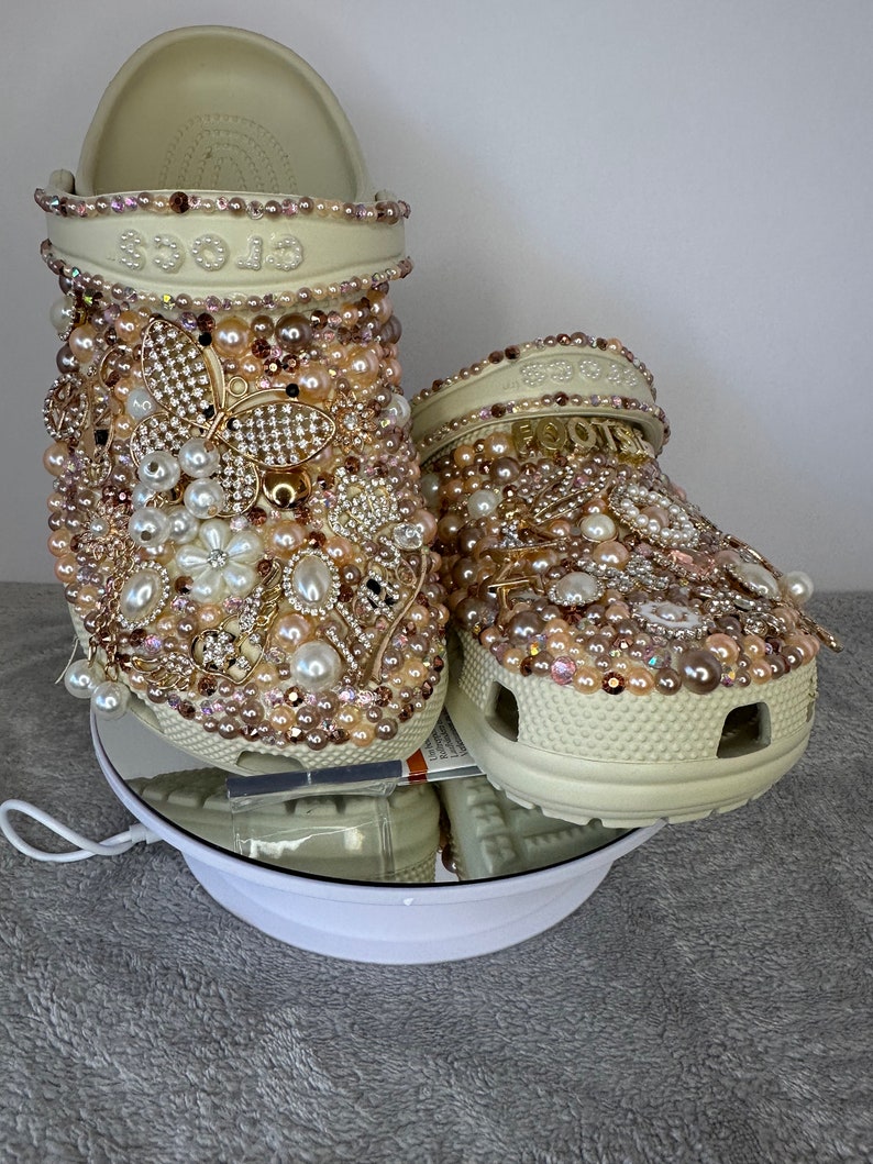 Pu&ograve; includere: Un paio di zoccoli bianchi decorati con perle, strass e dettagli dorati. Gli zoccoli hanno un design floreale e la parola "crocs" &egrave; visibile sulla parte superiore di uno zoccolo.