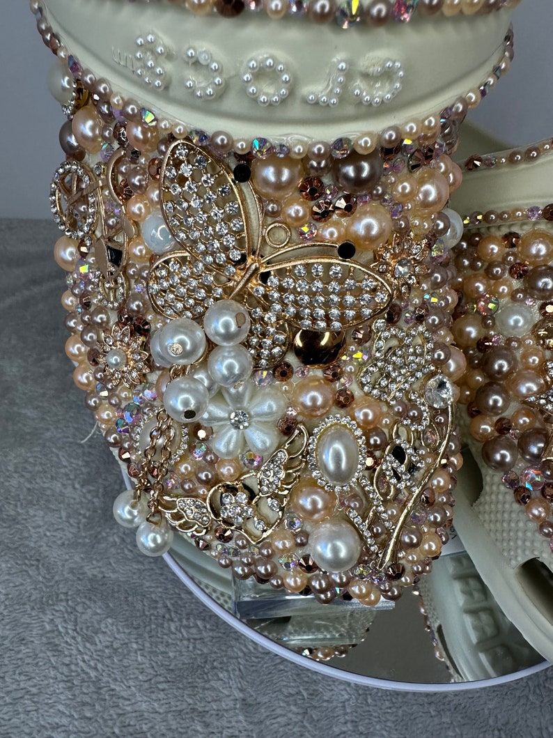 Pu&ograve; includere: Scarpa zoccolo bianca decorata con ornamenti dorati e perlati, tra cui un grande motivo a farfalla. La scarpa &egrave; su una superficie a specchio.