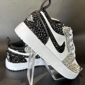 Puede incluir: Un par de zapatillas blancas con pedrer&iacute;a negra y transparente. Las zapatillas tienen un logotipo Swoosh negro y un dise&ntilde;o de pedrer&iacute;a negra y transparente en la puntera.