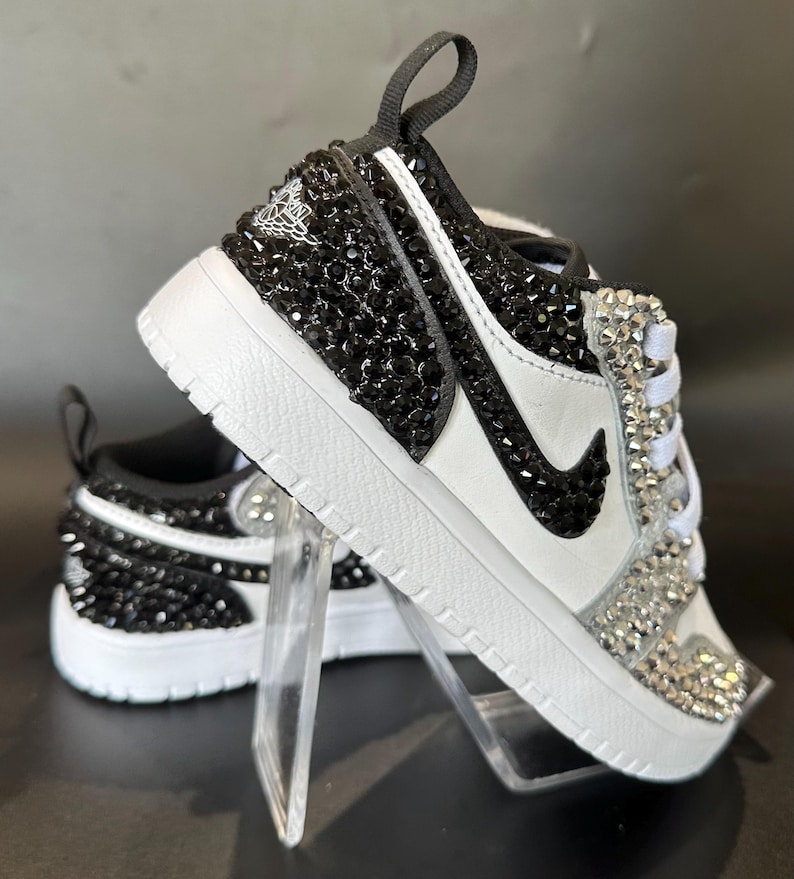 Puede incluir: Un par de zapatillas blancas con pedrer&iacute;a negra y transparente. Las zapatillas tienen un logotipo Swoosh negro y un dise&ntilde;o de pedrer&iacute;a negra y transparente en la puntera.