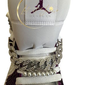 May include: Close-up of a white and purple sneaker with a silver chain and pearl embellishments. The tongue features a logo and the text "Let Her Designs". The laces are white.