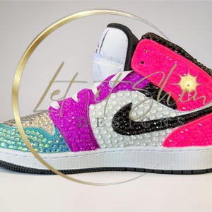 May include: A bedazzled high-top sneaker in vibrant colors. The shoe features a white base with a black swoosh, accented with pink, purple, and teal rhinestones. The design includes the text "Let Shine" and a starburst graphic.
