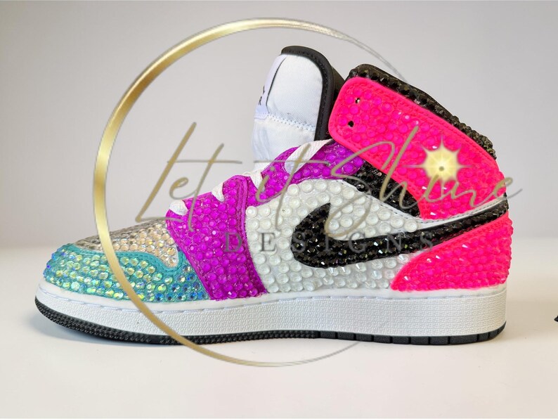 May include: A bedazzled high-top sneaker in vibrant colors. The shoe features a white base with a black swoosh, accented with pink, purple, and teal rhinestones. The design includes the text "Let Shine" and a starburst graphic.