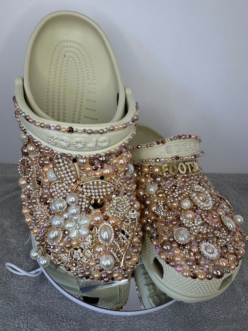 Pu&ograve; includere: Un paio di zoccoli bianchi decorati con perle, strass e dettagli dorati. Gli zoccoli sono impreziositi da un design di perle e strass e la parola "Footsie" &egrave; scritta in strass su uno zoccolo.