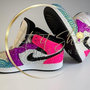 May include: Two bedazzled sneakers. One sneaker is primarily pink and purple with a black Nike swoosh. The other sneaker is primarily blue and white. Both sneakers have white soles and black laces. The sneakers are decorated with iridescent rhinestones.