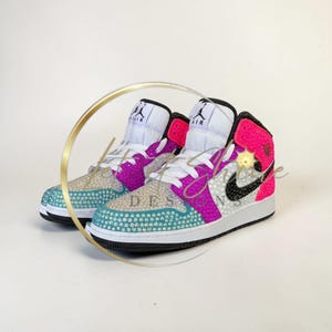 May include: A pair of customized high-top sneakers with a white, black, pink, and teal color scheme. The sneakers feature a jewel-encrusted design on the toe and side panels, with the Nike swoosh in black. The tongue is white with a black logo.