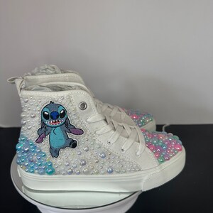 May include: White high-top sneakers with a blue Stitch character embroidered on the side. The shoes are covered in iridescent rhinestones.