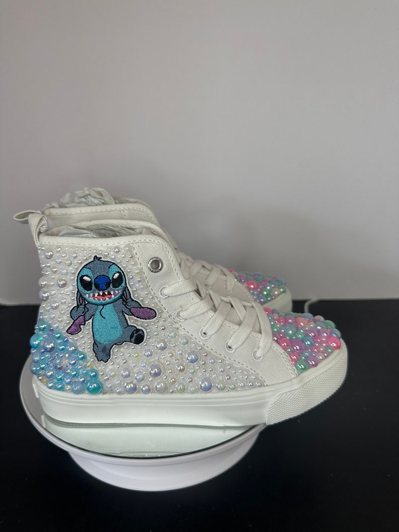 May include: White high-top sneakers with a blue Stitch character embroidered on the side. The shoes are covered in iridescent rhinestones.