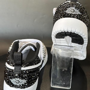 Puede incluir: Un par de zapatillas blancas con pedrer&iacute;a negra que cubre toda la zapatilla. Las zapatillas tienen un logotipo Air Jordan blanco y negro en el lateral.