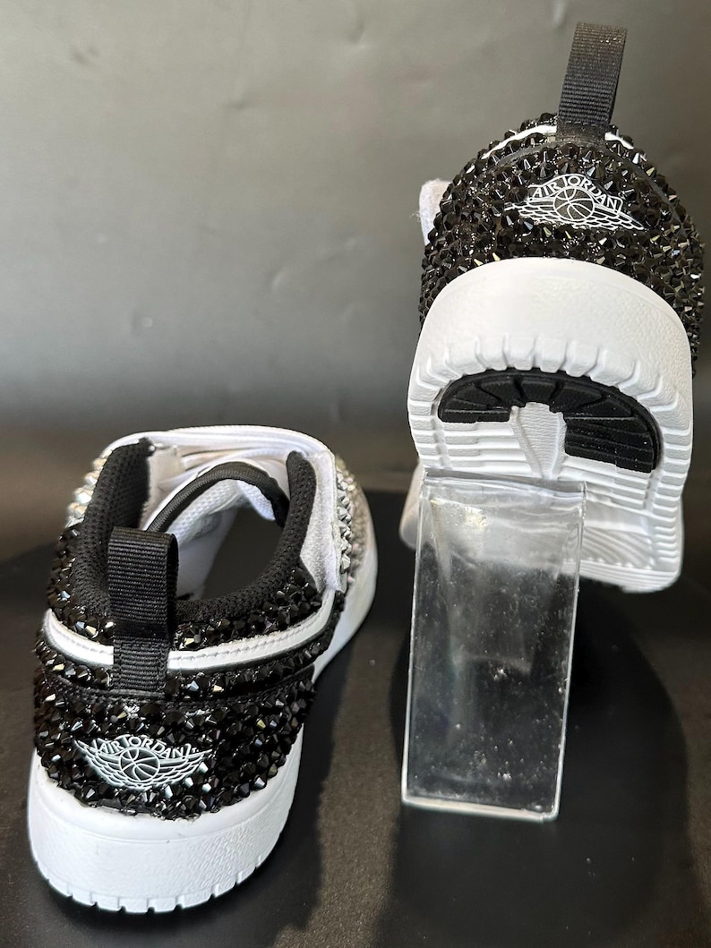 Puede incluir: Un par de zapatillas blancas con pedrer&iacute;a negra que cubre toda la zapatilla. Las zapatillas tienen un logotipo Air Jordan blanco y negro en el lateral.