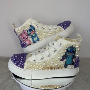 May include: White high-top sneakers with purple and white pearl embellishments. The sneakers feature an embroidered image of Stitch and Angel from the Disney movie Lilo & Stitch. The name "COURTLYN" is written in gold lettering on the side of the shoe.
