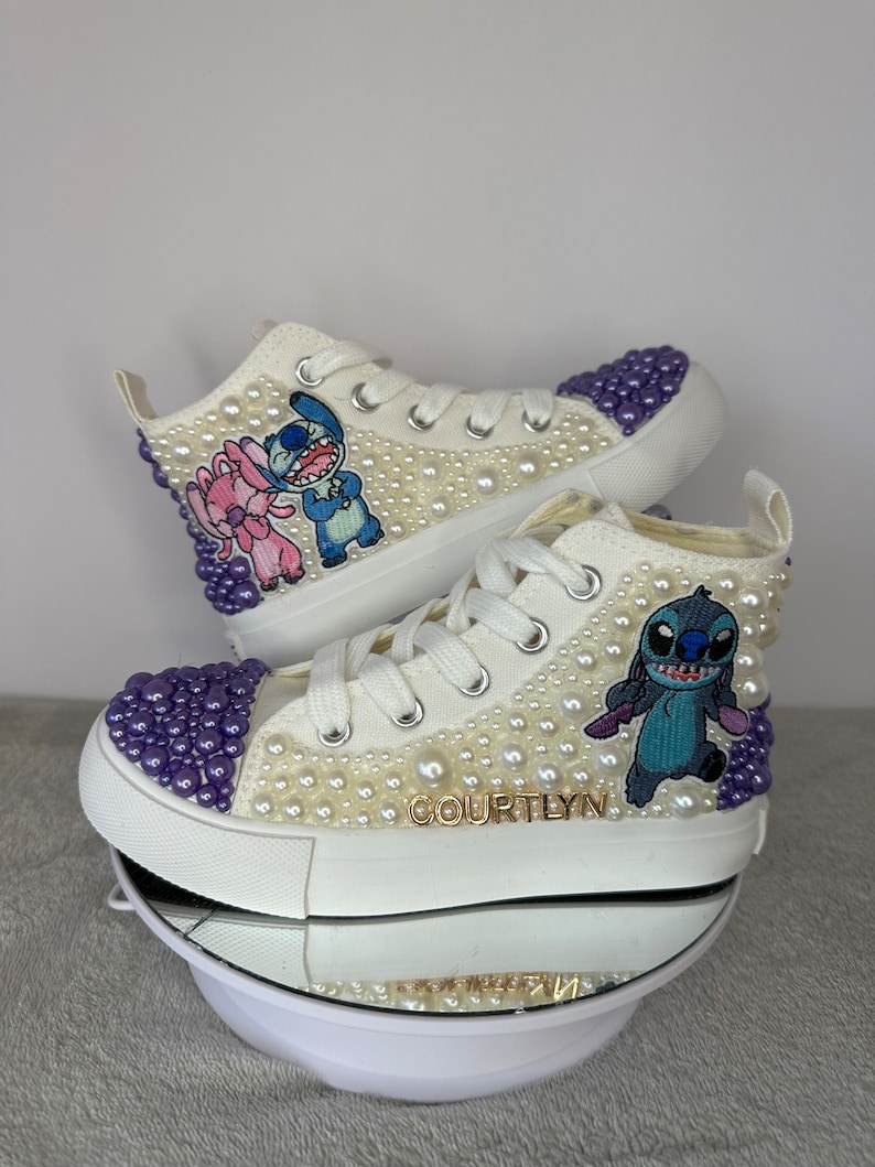 May include: White high-top sneakers with purple and white pearl embellishments. The sneakers feature an embroidered image of Stitch and Angel from the Disney movie Lilo & Stitch. The name "COURTLYN" is written in gold lettering on the side of the shoe.
