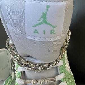 May include: White high-top sneakers with a green "AIR" logo and a silver chain around the ankle. The shoes are decorated with rhinestones and pearls.