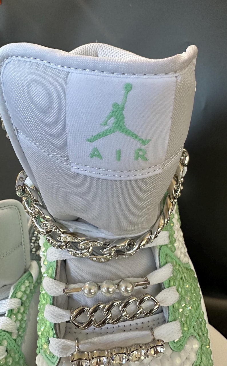 May include: White high-top sneakers with a green "AIR" logo and a silver chain around the ankle. The shoes are decorated with rhinestones and pearls.