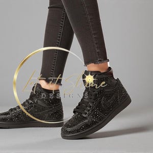 May include: Black high-top sneakers with sparkling embellishments. The shoes feature a lace-up design and a black sole. The sneakers are worn with black denim jeans. The overall aesthetic is stylish and eye-catching.