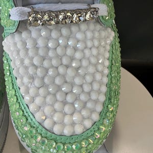 May include: Close-up of a white sneaker with a green trim. The sneaker is covered in white and green rhinestones. A silver chain and a row of clear rhinestones are on the laces.